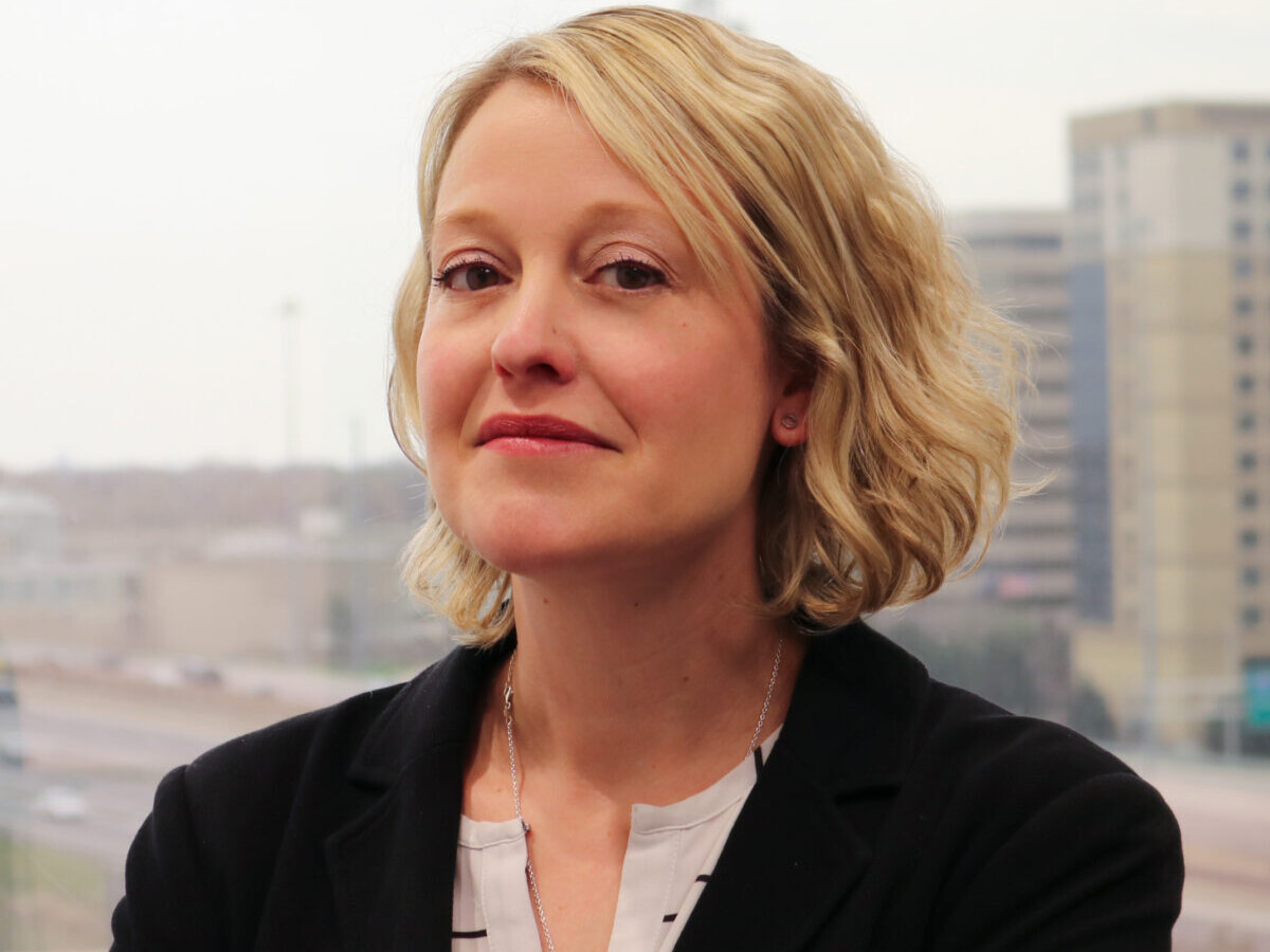 A woman in black jacket and white shirt near window.